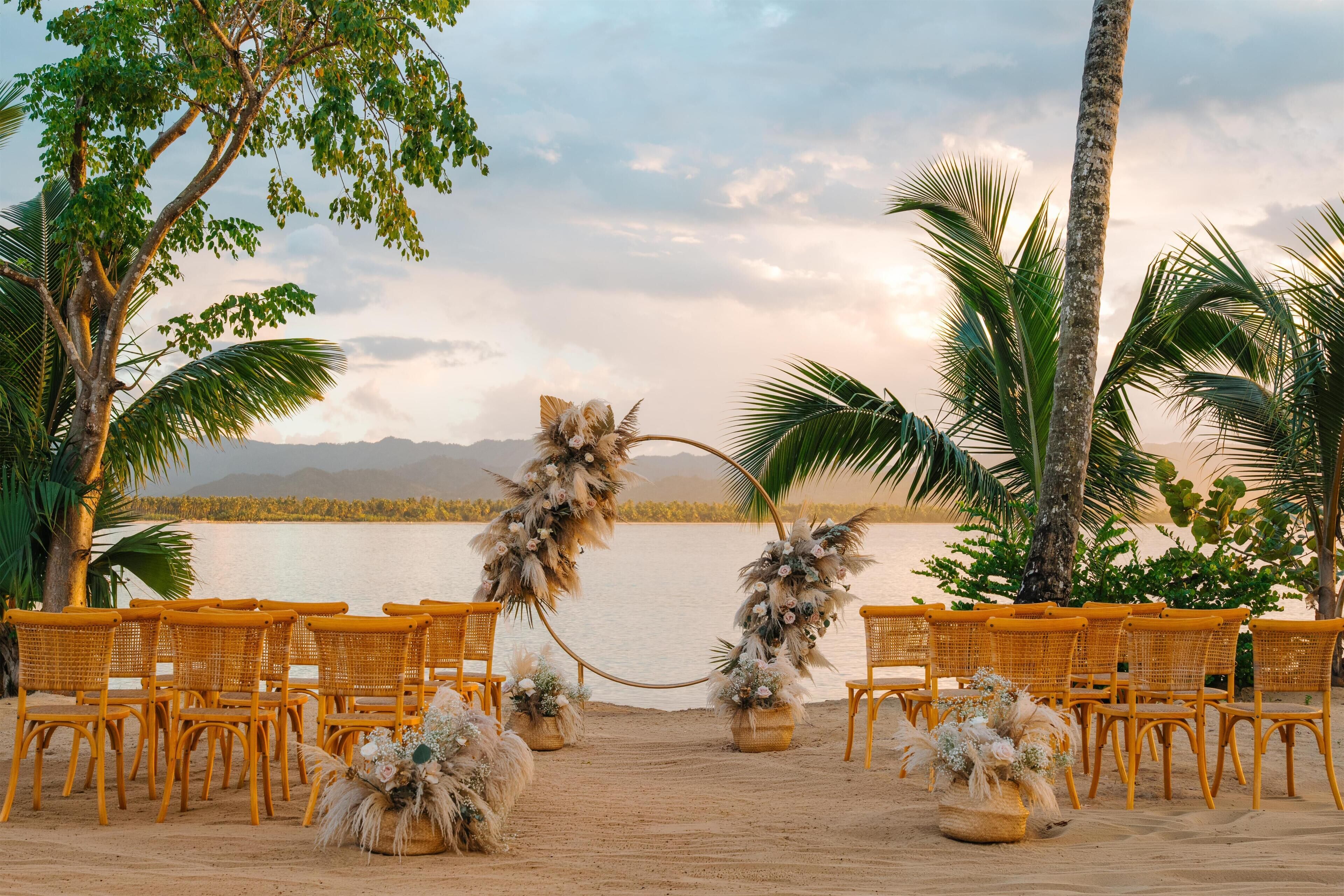 A beach setting with wicker chairs arranged in rows, a decorative archway, and a backdrop of palm trees and a body of water.