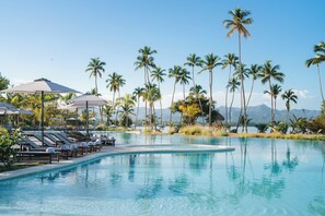 Piscina com espreguiçadeiras e guarda-sóis, cercada por palmeiras e montanhas.