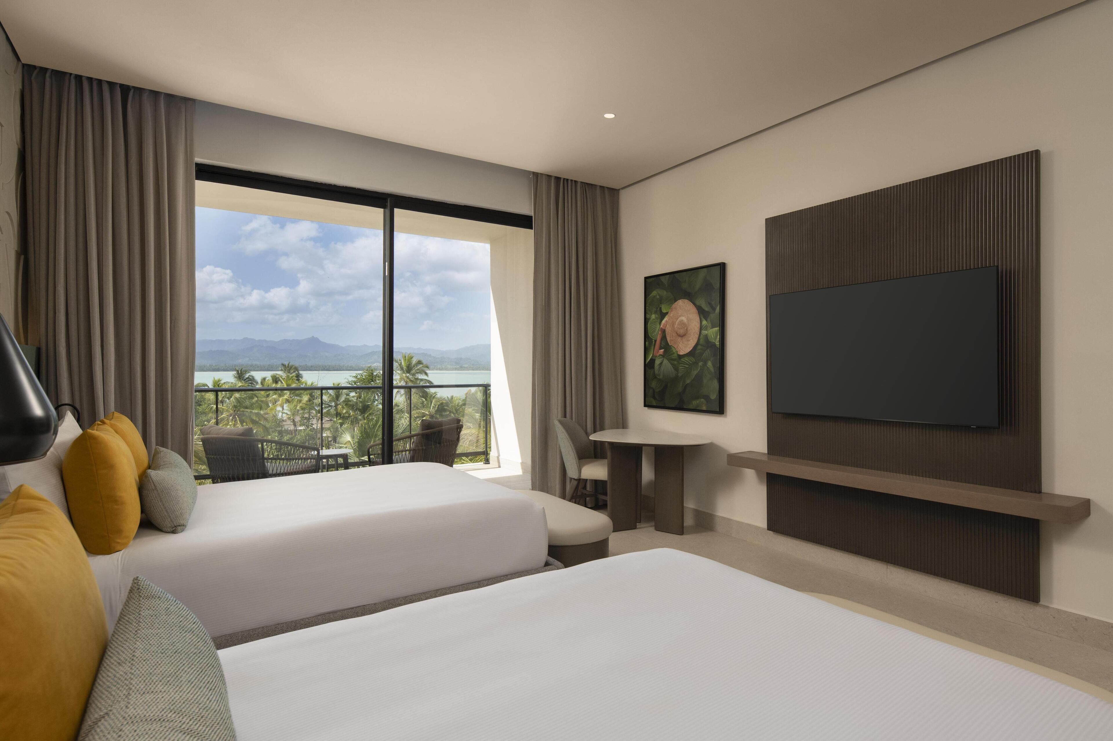 Quarto de hotel com duas camas, uma mesa, uma cadeira, uma TV e uma grande janela com vista para palmeiras e montanhas.