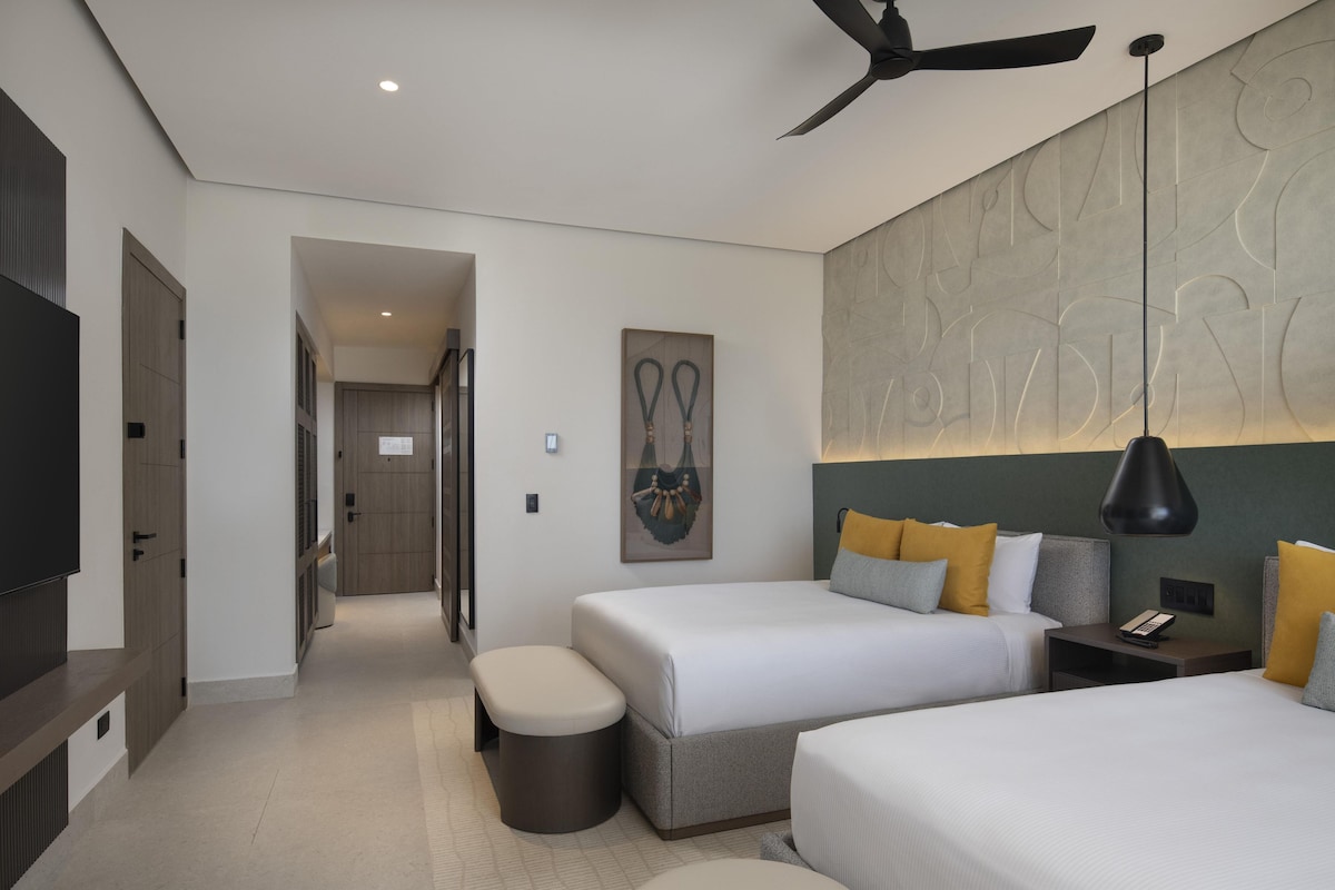 A modern hotel room with two beds, a ceiling fan, a flat-screen TV, and a large abstract wall art piece.