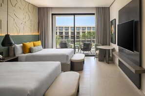Quarto de hotel com duas camas, televisão, mesa pequena e varanda com vista para edifícios e palmeiras.