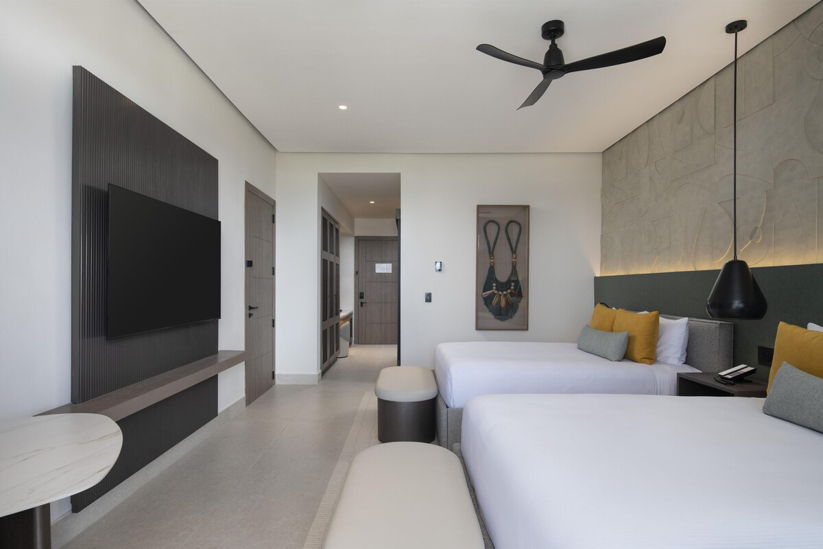 Quarto de hotel moderno com televisão de tela plana, duas camas, ventilador de teto e uma grande obra de arte na parede.