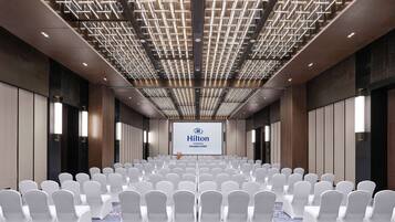 A conference room with rows of white chairs, a blue carpet, and a ceiling with a geometric light fixture. A Hilton logo is visible on the wall.