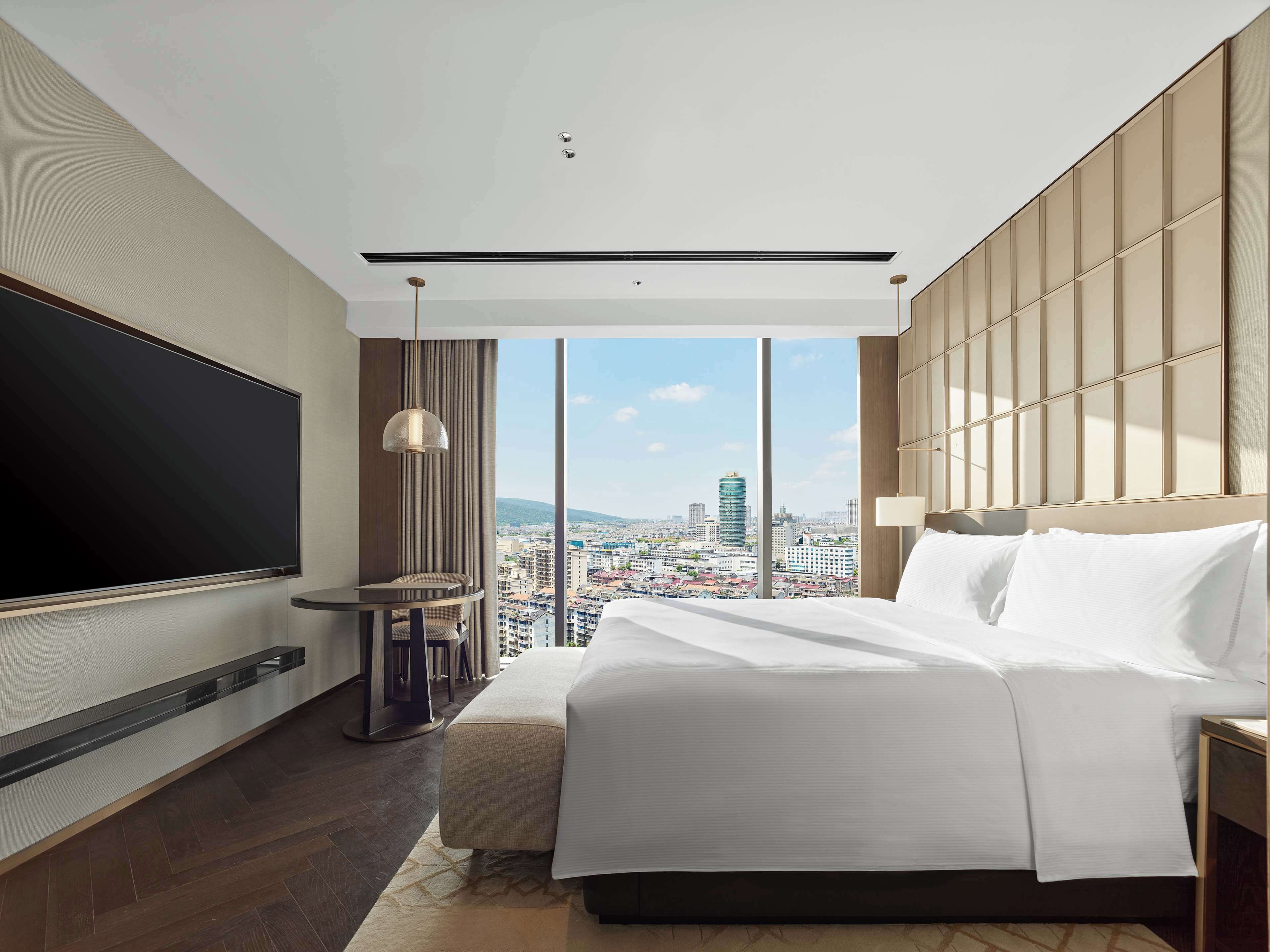 A modern hotel room with a large bed, a flat-screen TV, a small table, and a city view.