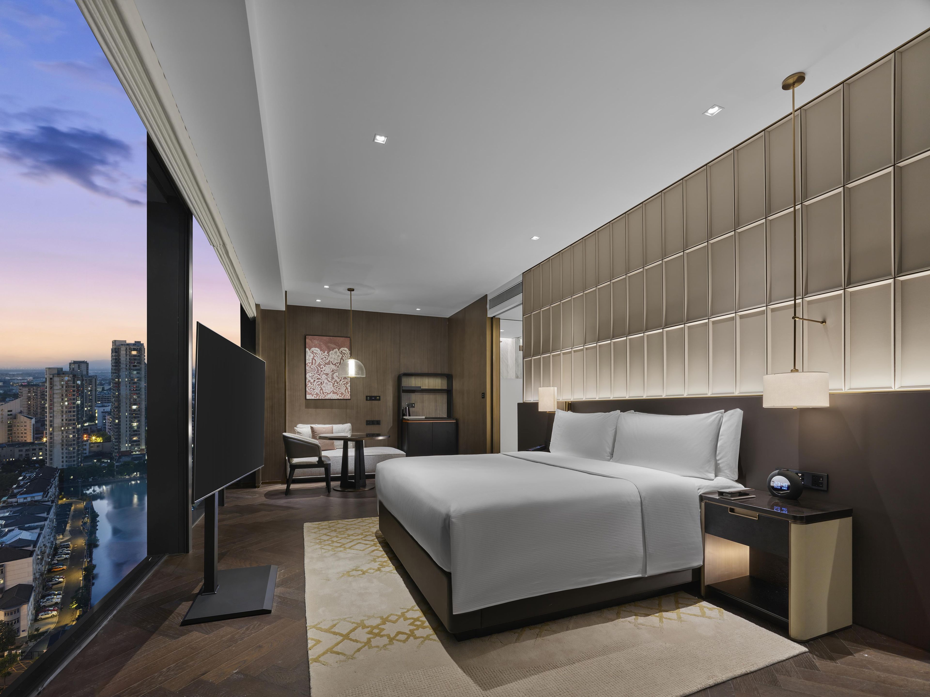 A modern hotel room with a large bed, bedside tables, a TV, and a city view.