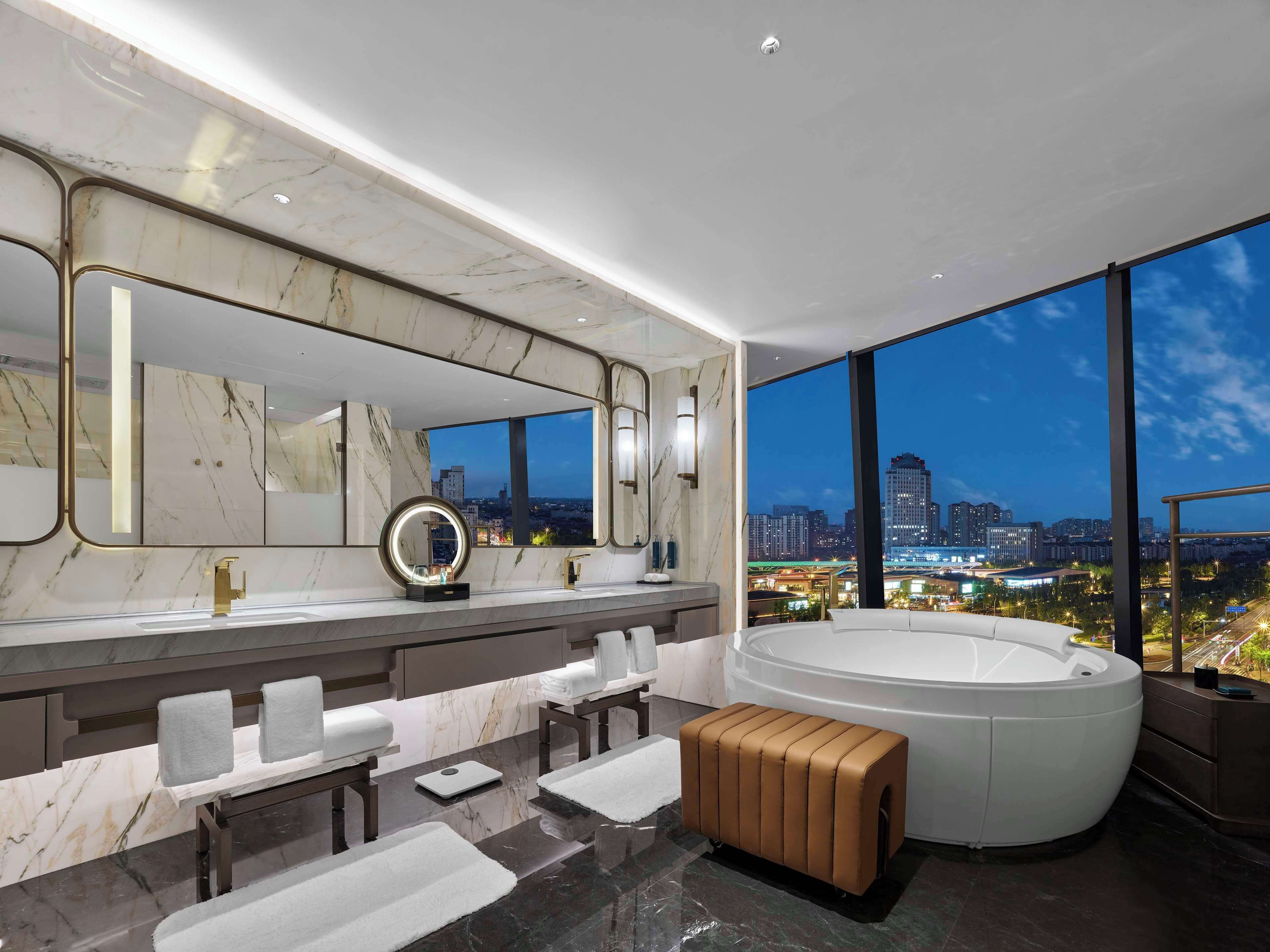 A modern bathroom with a large mirror, two sinks, a bathtub, and a view of a cityscape at night.