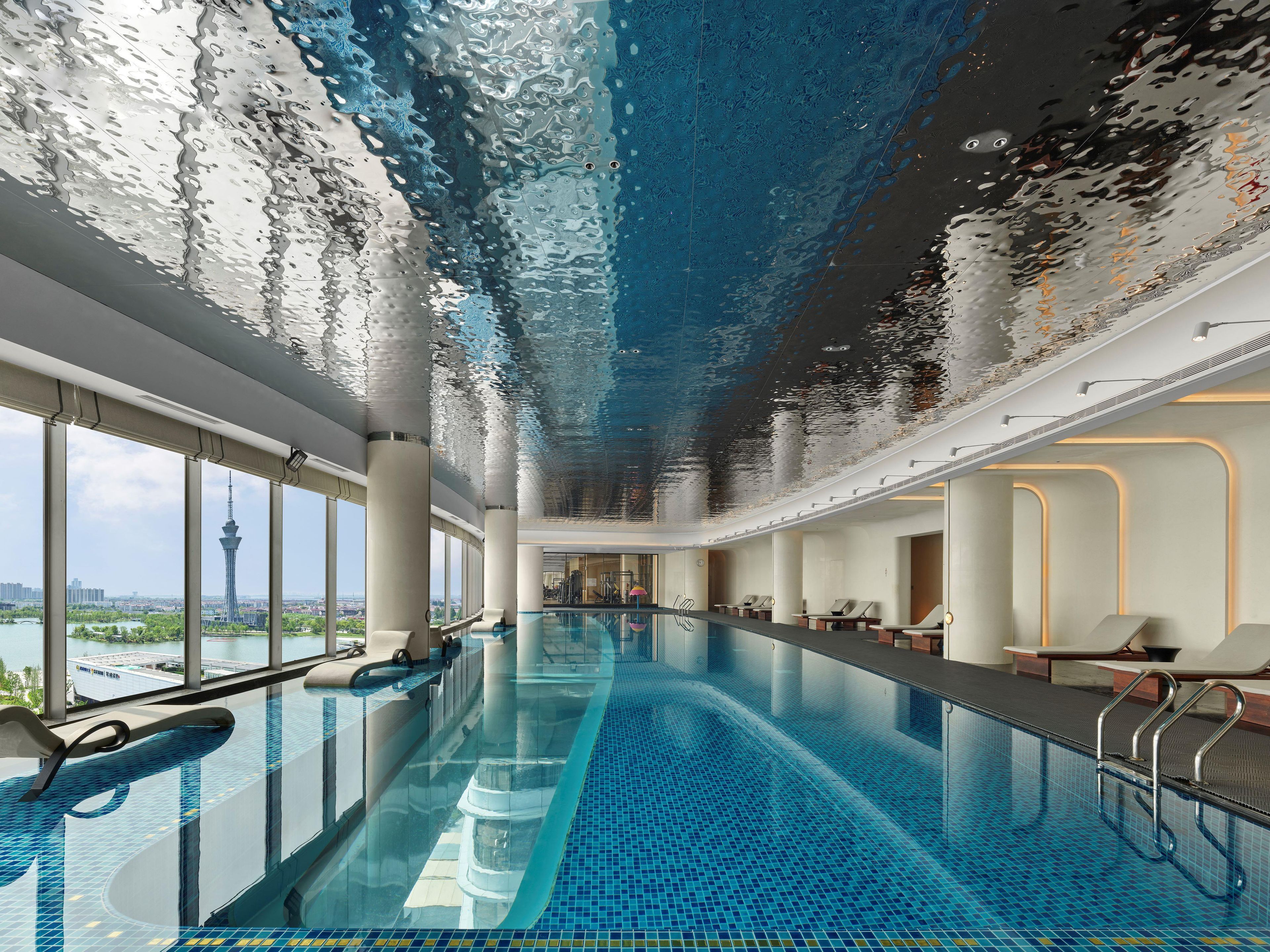 Una piscina interior moderna con paredes de vidrio transparente, vista a un paisaje urbano y suelo de baldosas.