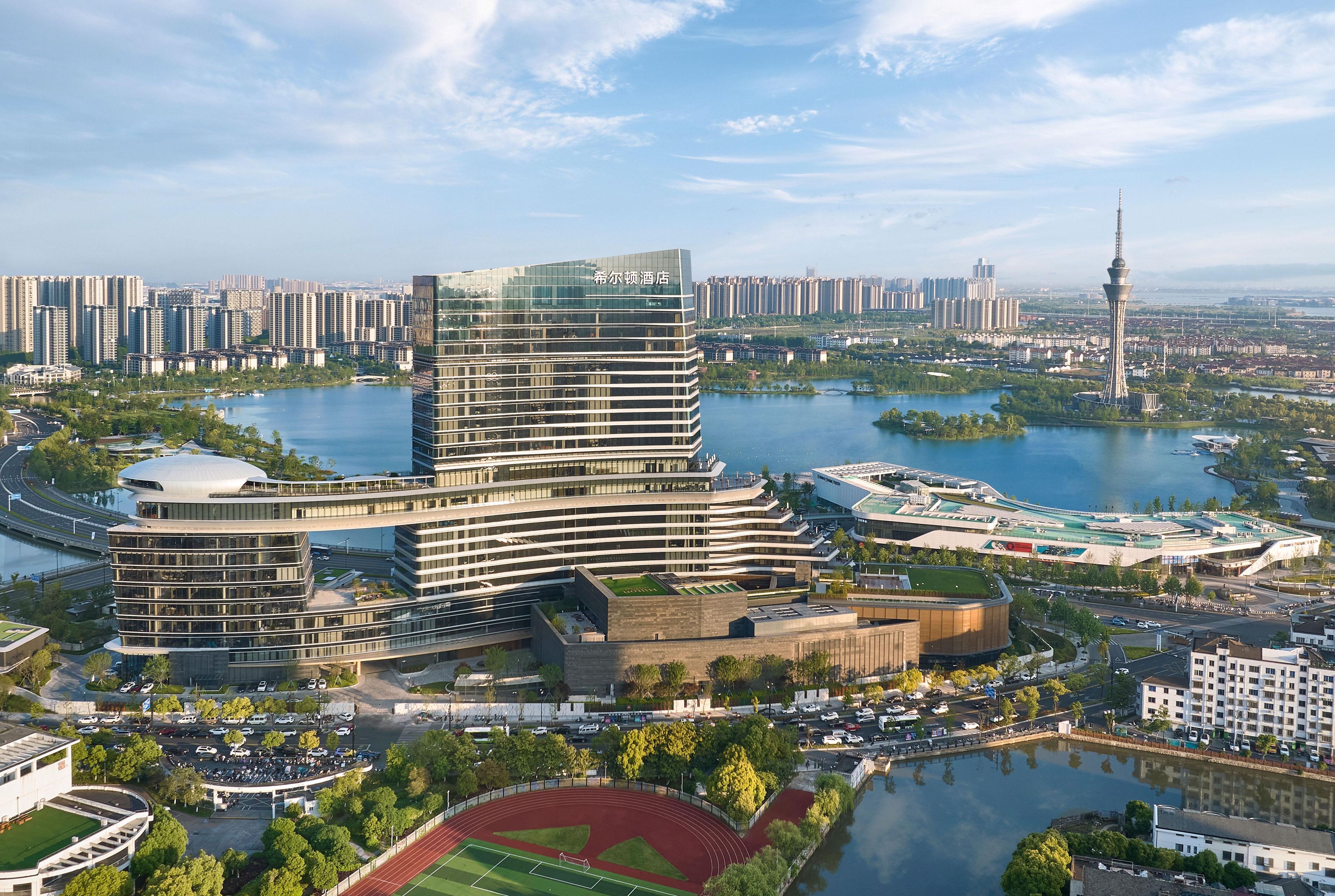 A modern hotel with a unique curved architecture, situated near a body of water and surrounded by a cityscape.
