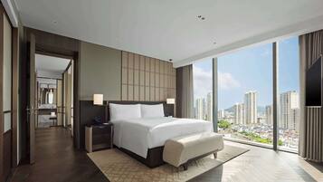 A modern hotel room with a large bed, bedside lamps, a view of the cityscape, and a walk-in closet.