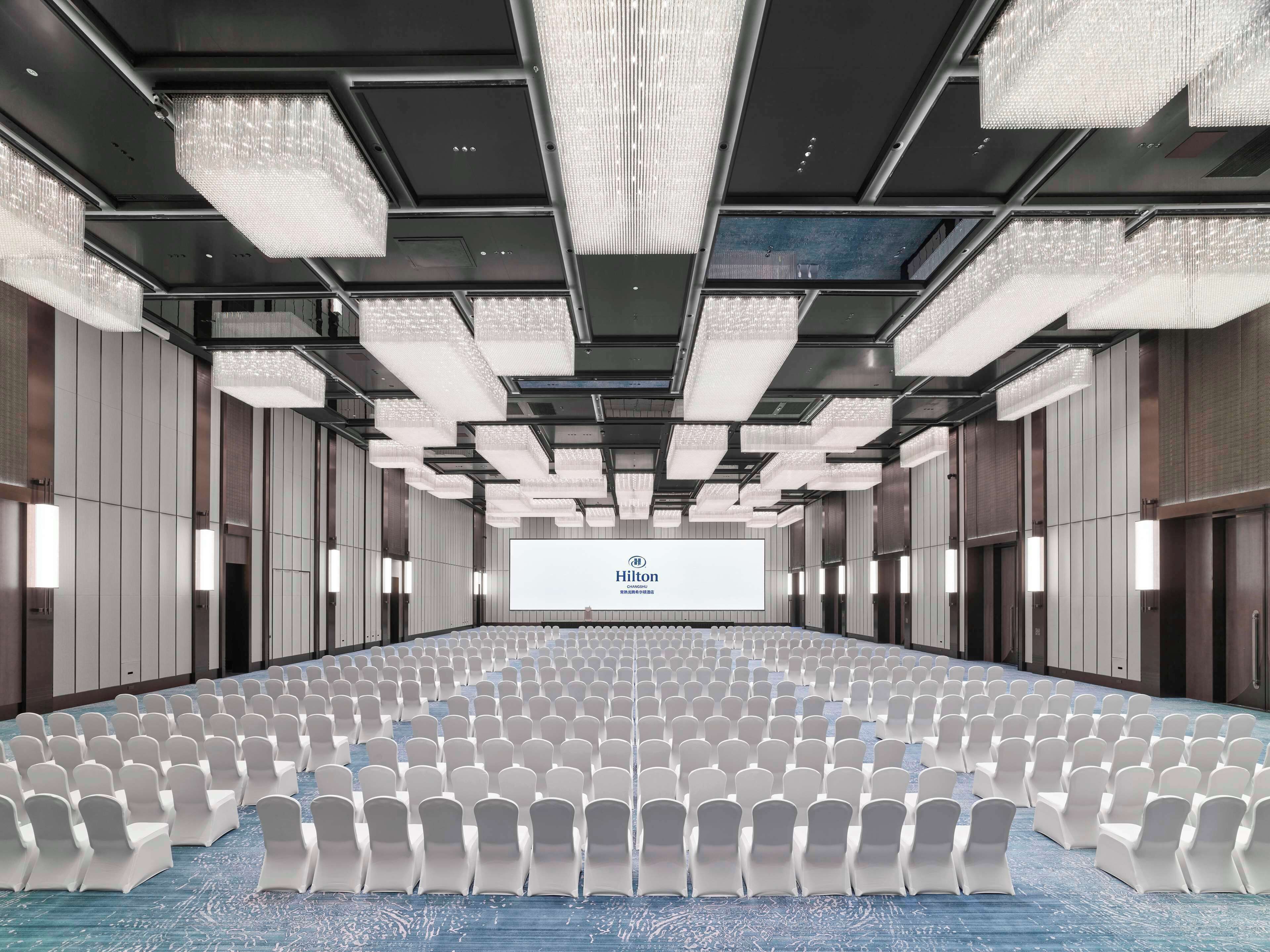 A large conference room with rows of white chairs, a large screen at the front, and modern chandeliers.