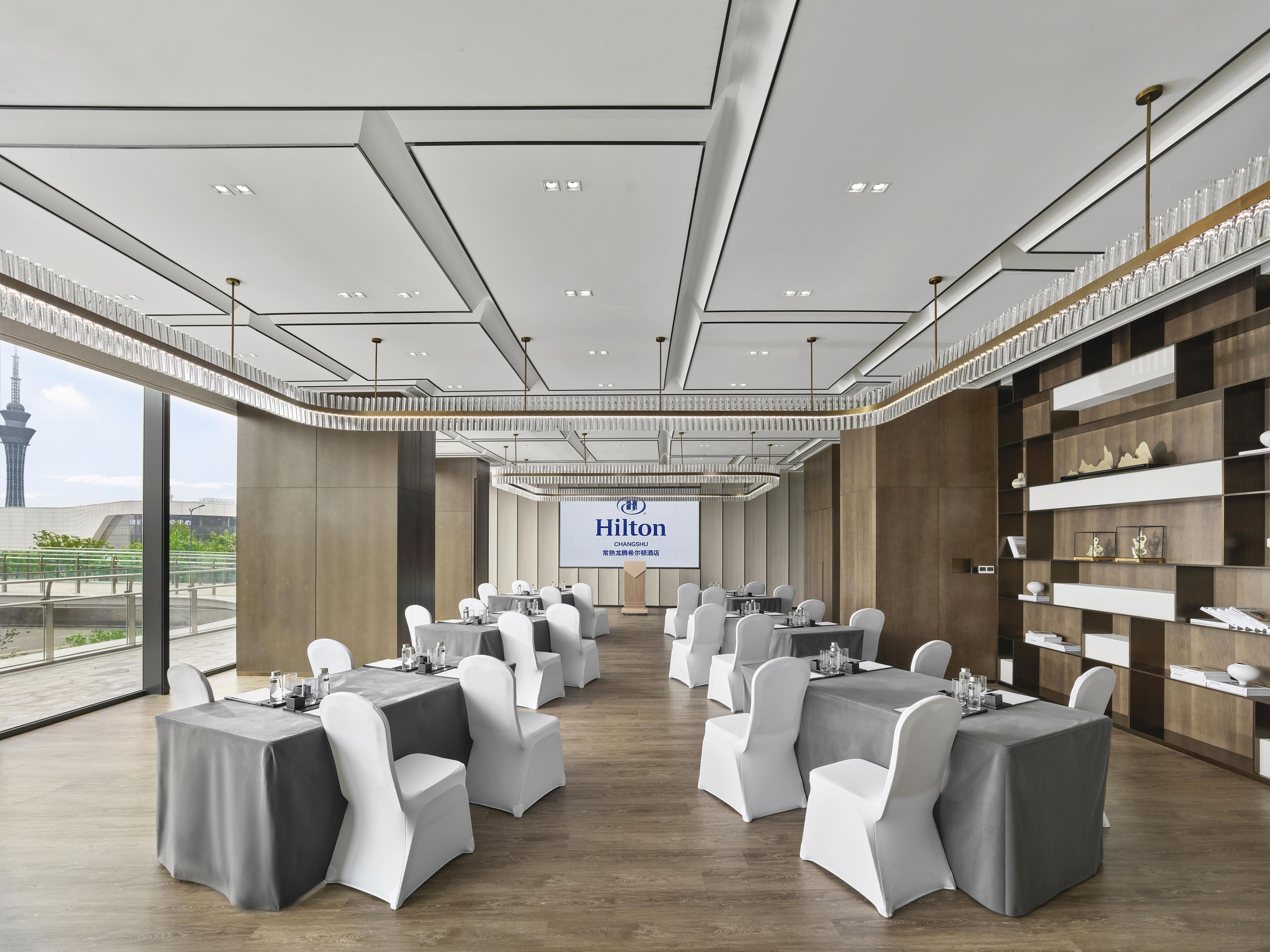 A conference room with multiple tables and chairs, a large screen displaying the Hilton logo, and a view of a tower through large windows.