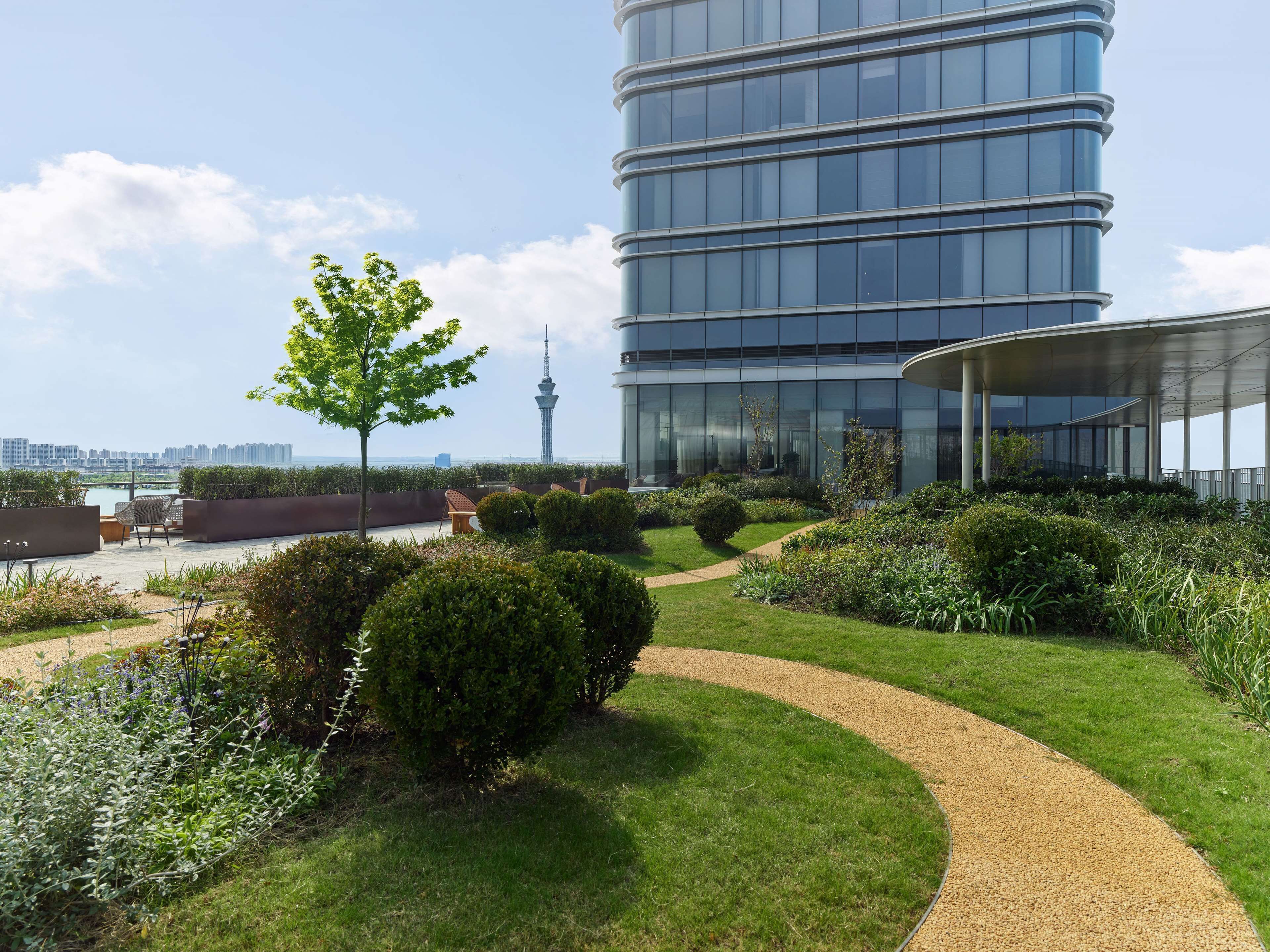 A modern building with a curved glass facade, a rooftop garden with pathways, and a view of a tower in the distance.