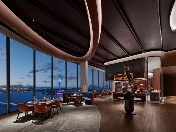 A modern interior with a panoramic view of a city skyline and a tower, featuring a bar area and seating arrangements. - Hilton Changshu (Suzhou)