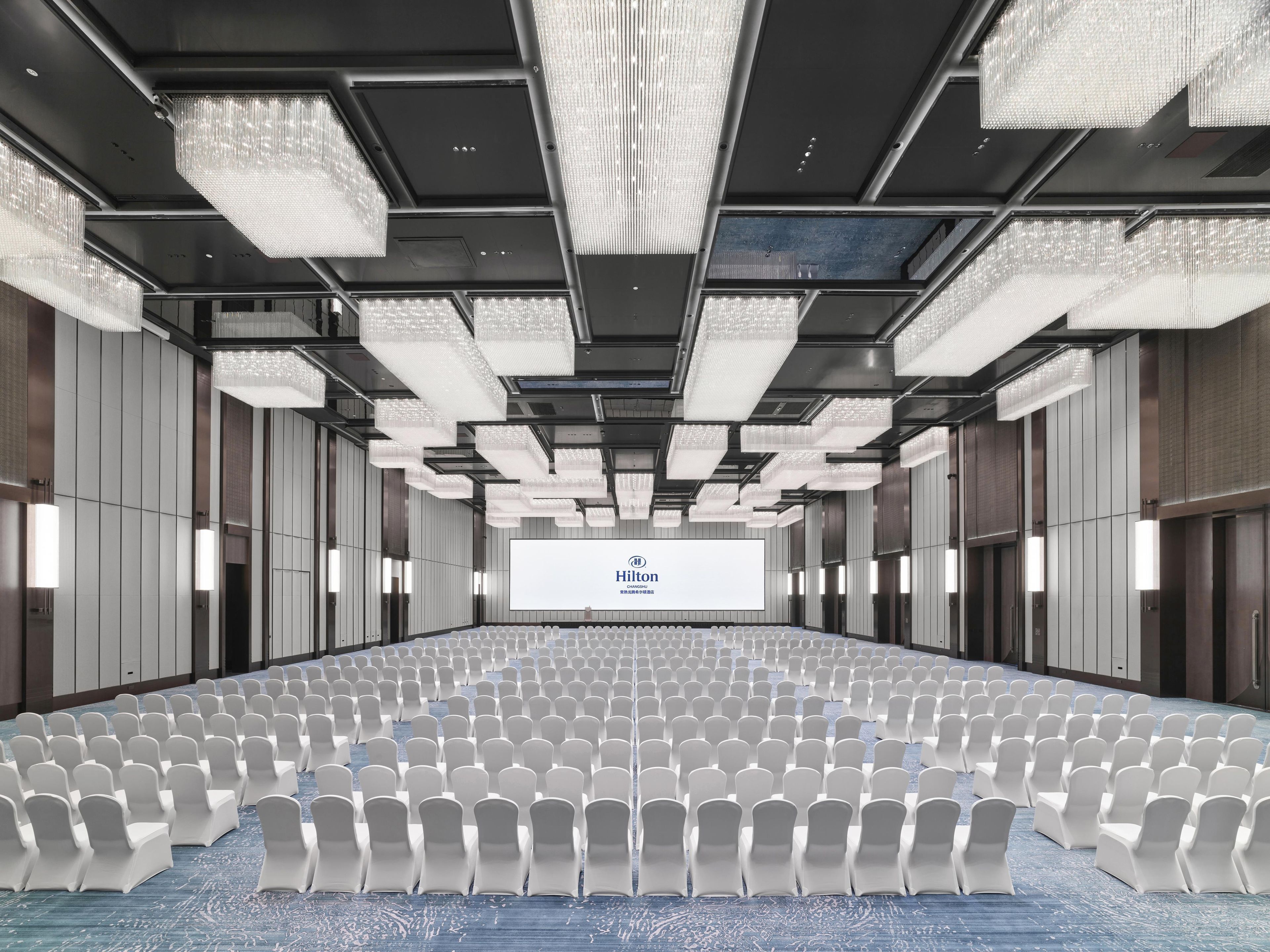 A conference room with rows of white chairs, a large screen at the front, and modern chandeliers.