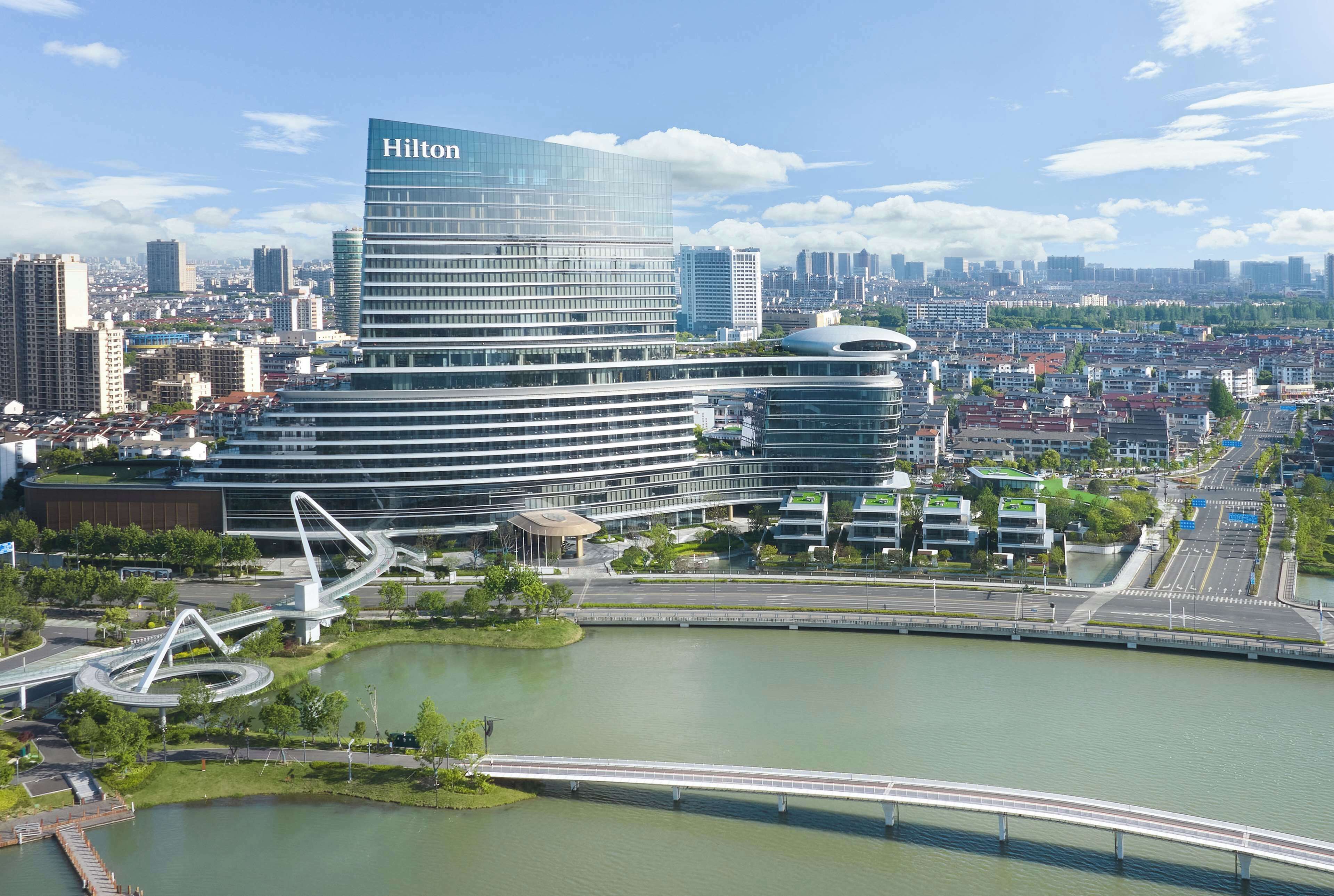 Un hotel moderno con un llamativo letrero de Hilton, rodeado por un paisaje urbano y una masa de agua.
