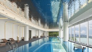 An indoor swimming pool with clear blue water, a glass ceiling reflecting the sky, and a cityscape view through large windows. - Hilton Changshu (Suzhou)