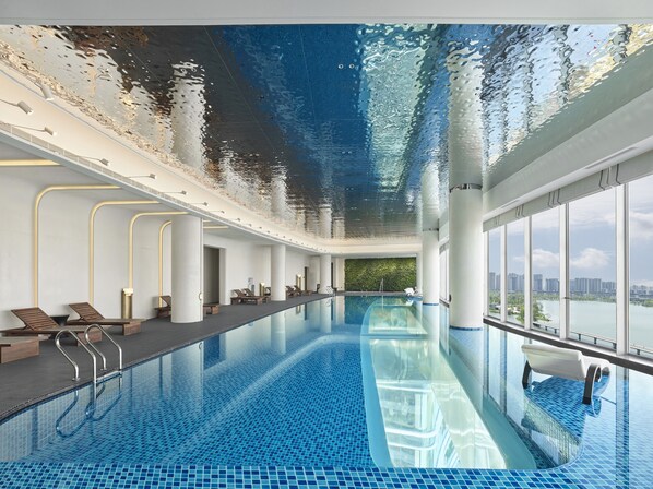 An indoor swimming pool with clear blue water, a glass ceiling reflecting the sky, and a cityscape view through large windows. - Hilton Changshu (Suzhou)