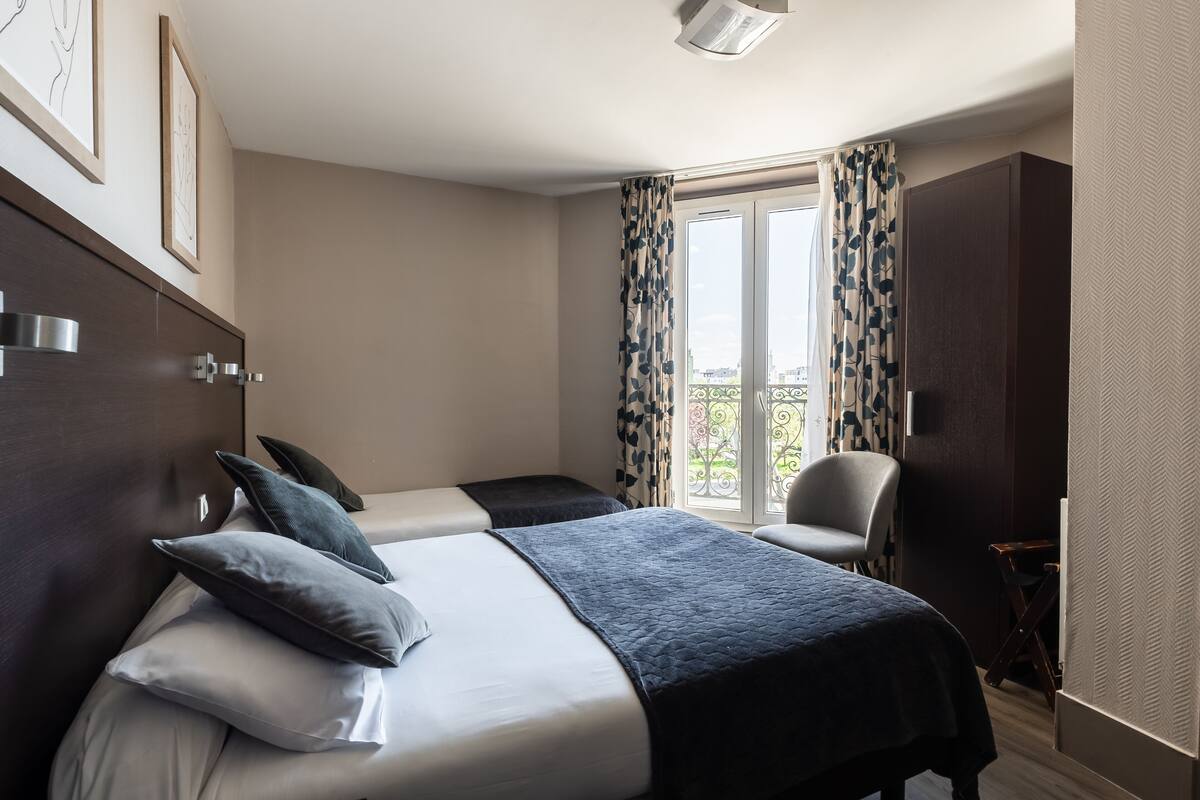 A hotel room with two beds, a chair, a wardrobe, and a balcony with curtains.