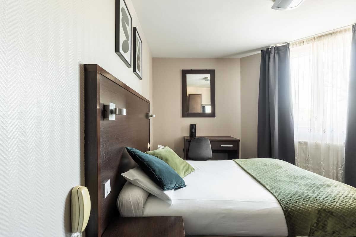 A hotel room with a bed, a desk, a chair, a mirror, and a window with curtains.