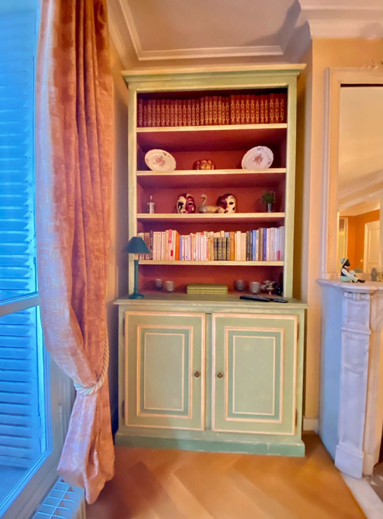 A room with a green bookshelf filled with books and decorative items, a window with blinds, and a door leading to another room.