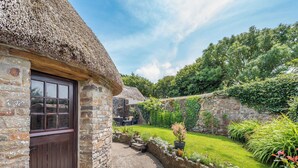 A stone cottage with a thatched roof, a wooden door, and a stone pathway leading to a garden with various plants and a stone wall. - East Titchberry Cottage (Hartland)