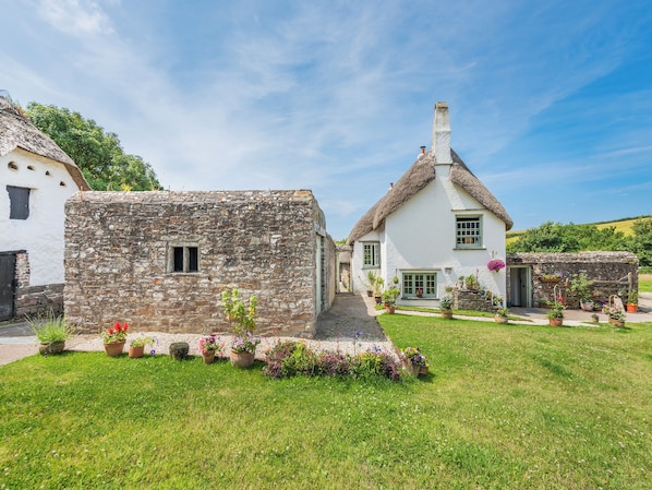 A traditional stone building with a thatched roof, a white house with green shutters, and a well-maintained garden with various potted plants.