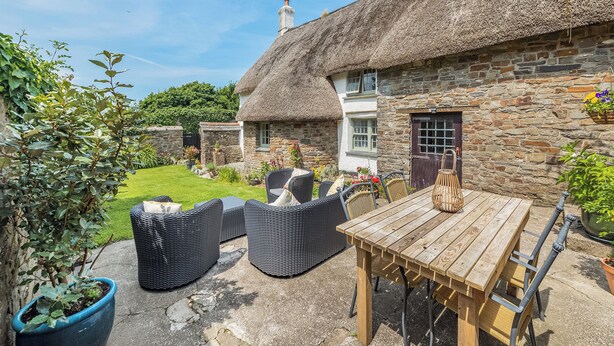 A thatched-roof cottage with a stone exterior, a wooden table, wicker chairs, and a potted plant. - East Titchberry Cottage (Hartland)