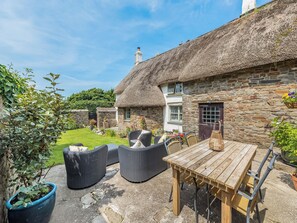 A thatched-roof cottage with a stone exterior, a wooden table, wicker chairs, and a potted plant.