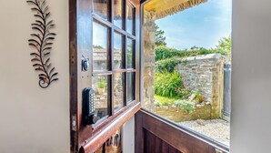 A wooden door with a glass panel, a metal decorative piece on the wall, and a view of a stone wall and greenery outside. - East Titchberry Cottage (Hartland)