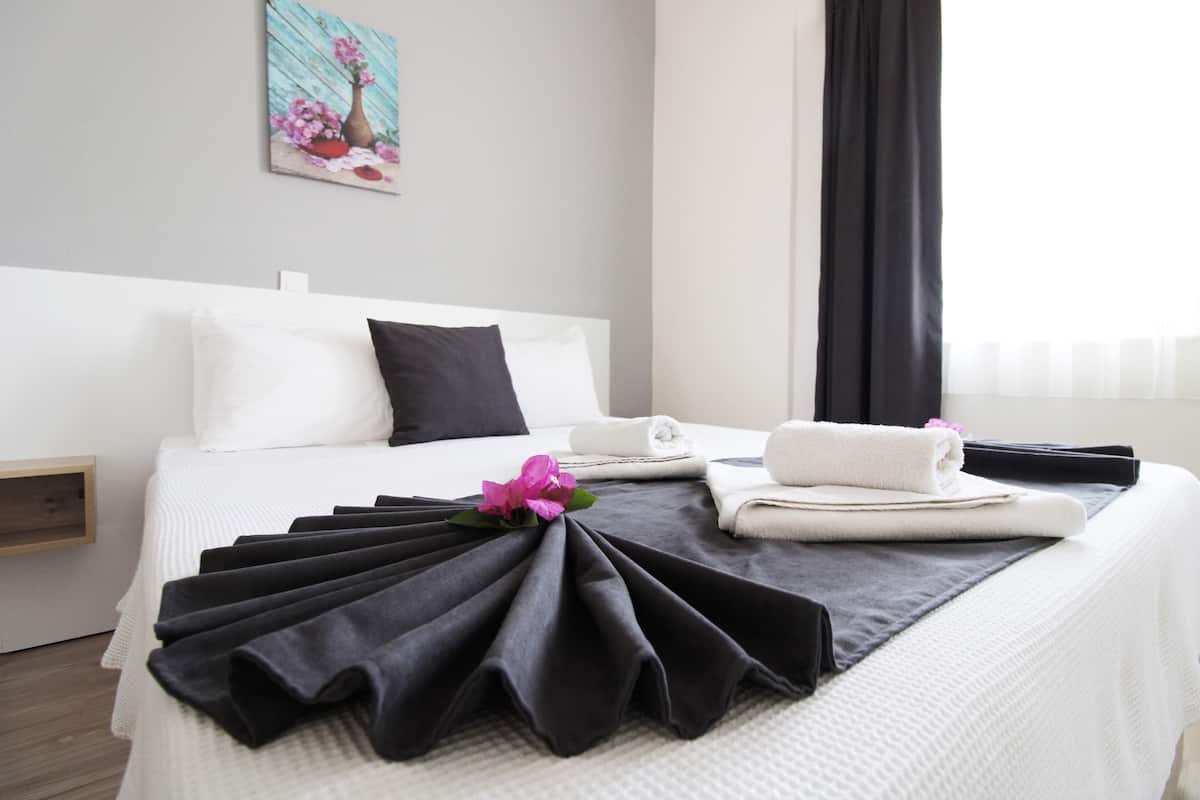 Un lit bien fait, agrémenté d’une serviette noire pliée en éventail, de coussins blancs et d’un petit vase avec des fleurs roses placé sur le mur.