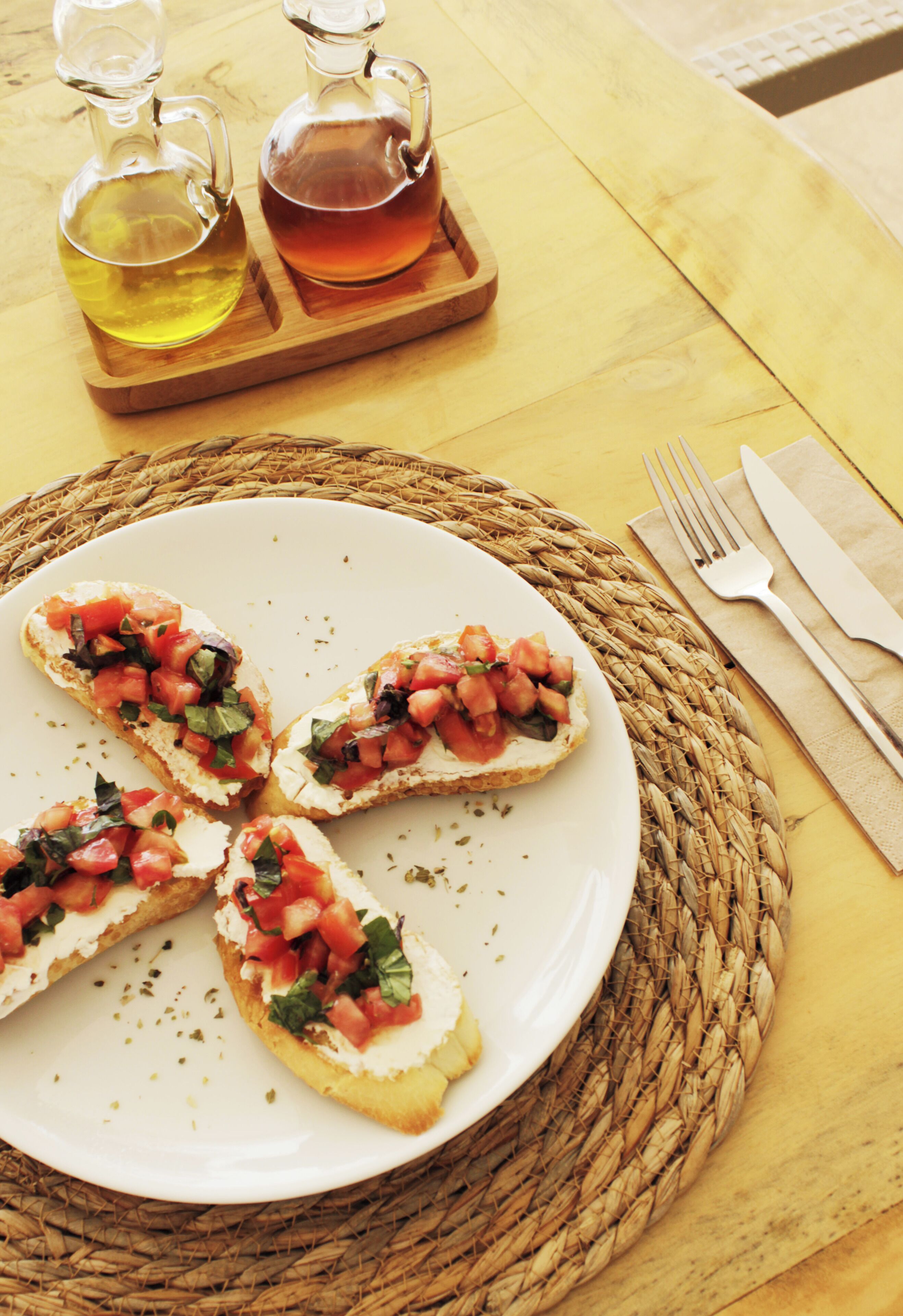 A plate of bruschetta with tomatoes and basil, a fork, and a knife on a table with a woven placemat.