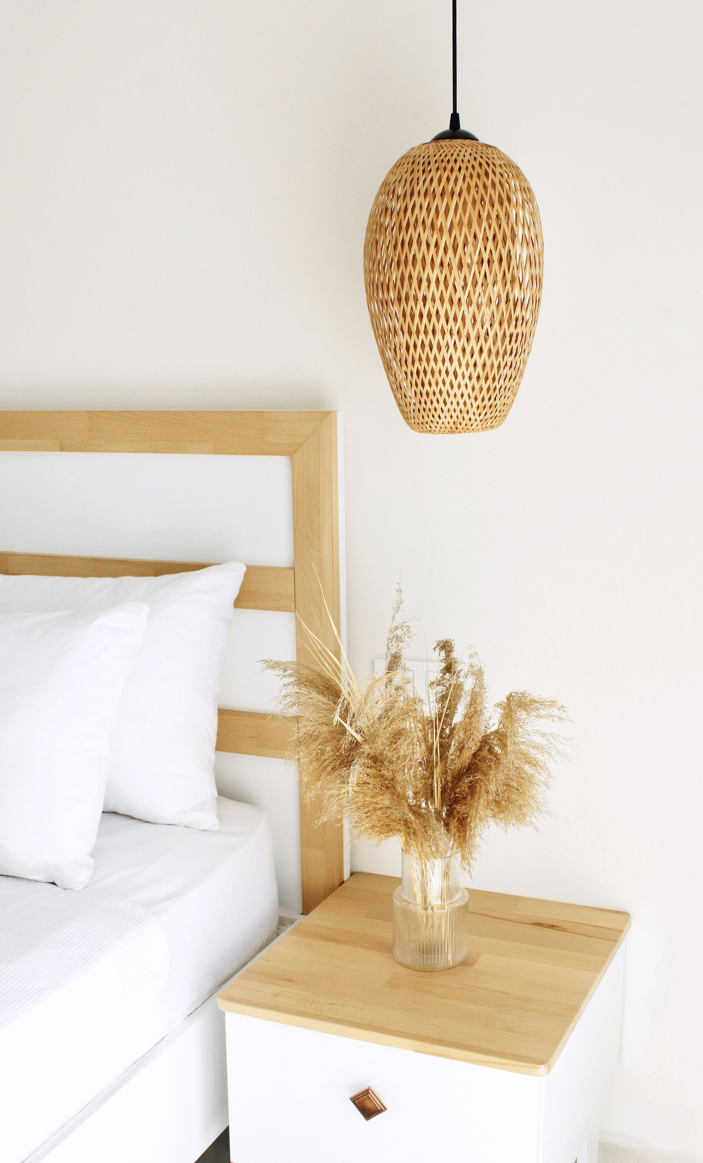 A bedroom with a wooden headboard, white bedding, a bedside table with a vase of dried plants, and a woven pendant light.