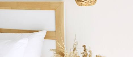 A bedroom with a wooden headboard, white bedding, a bedside table with a vase of dried plants, and a woven pendant light.