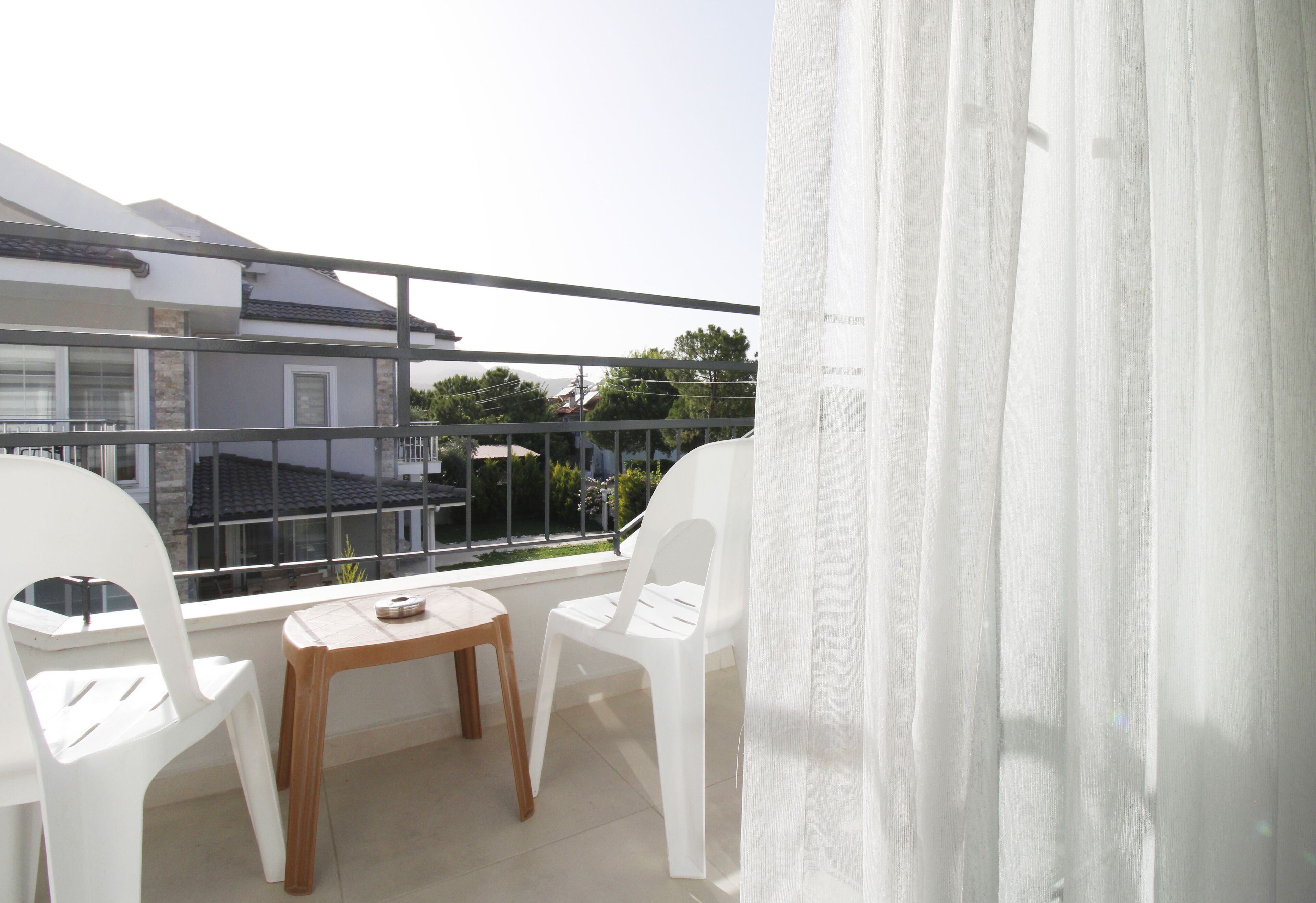 A balcony with white plastic chairs, a wooden table, and a view of buildings and trees.