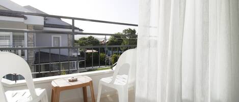 A balcony with white plastic chairs, a wooden table, and a view of buildings and trees.