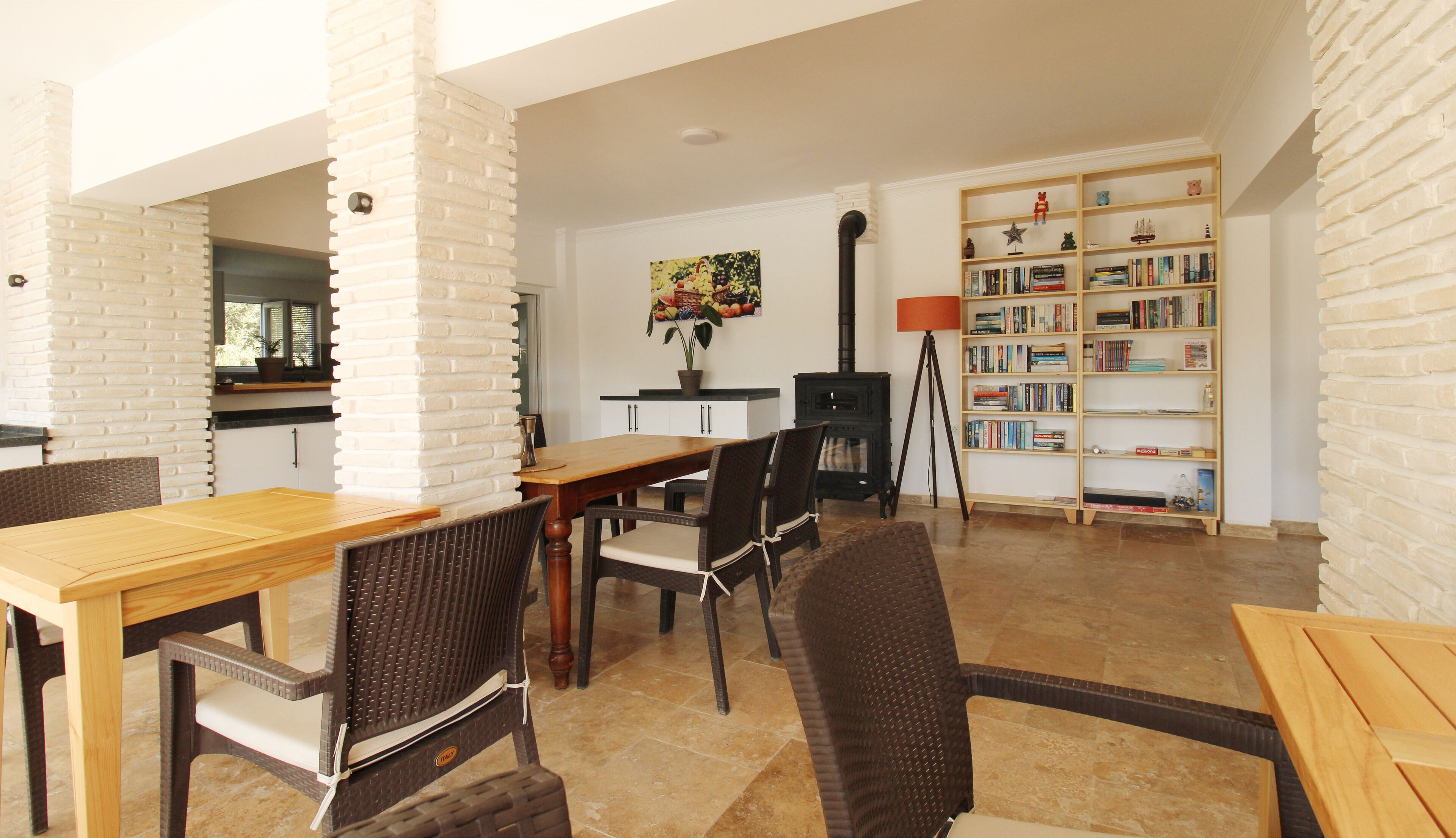 A dining area with a wooden table and wicker chairs, a bookshelf with books, and a decorative wall piece.