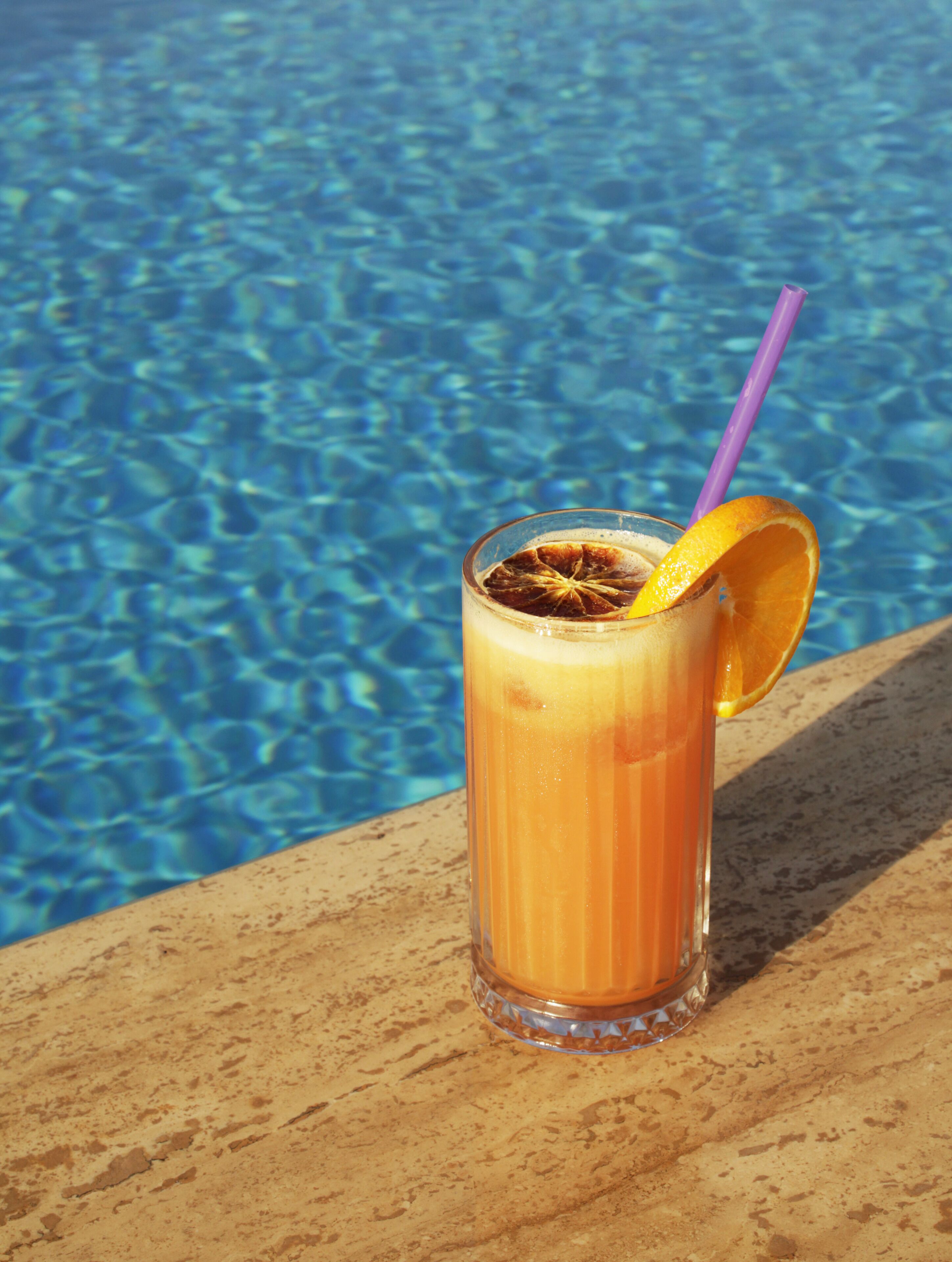 Un vaso de bebida anaranjada con una pajita morada y una rodaja de naranja, sobre una superficie de madera junto a una piscina.