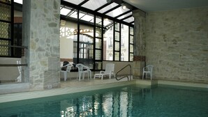 Una piscina interior con agua cristalina, paredes de piedra y sillas blancas.