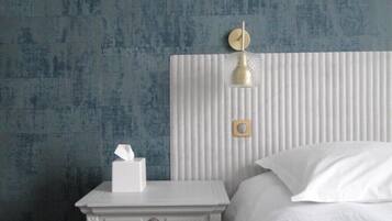 A neatly made bed with white linens, a white bedside table with a tissue box, and a wall-mounted light fixture.