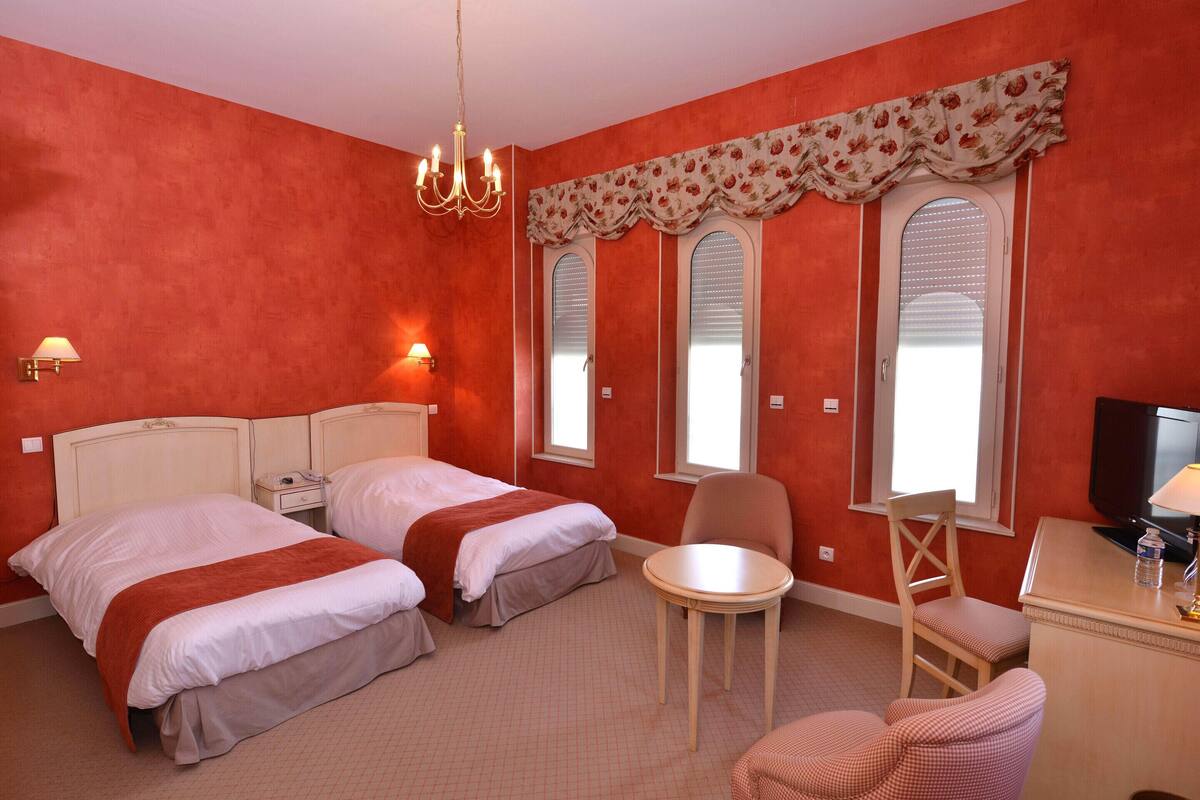 Ein Hotelzimmer mit zwei Betten, einem Sessel, einem kleinen Tisch, einem Fernseher und einem Fenster mit Rollläden.