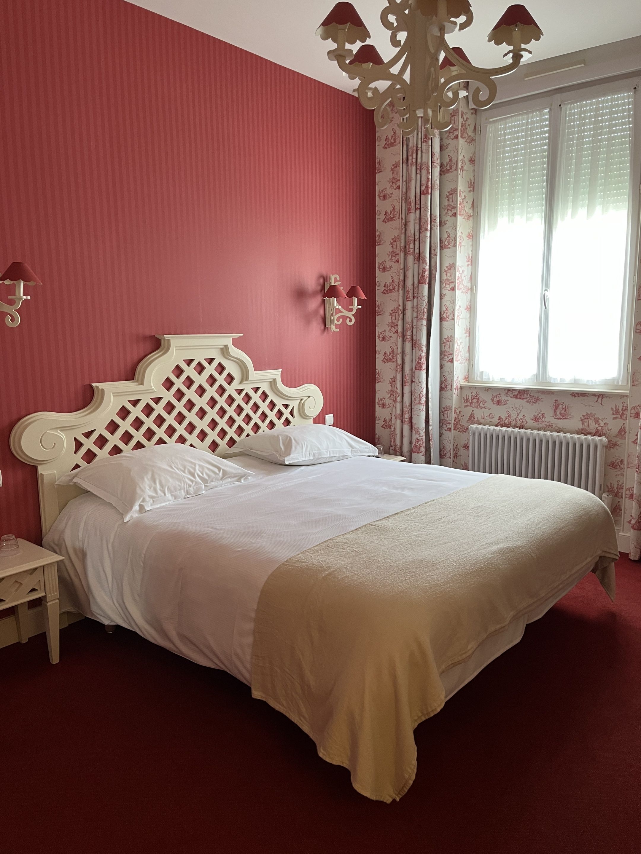 A bedroom with a bed, a nightstand, a decorative wall sconce, a window with curtains, and a radiator.
