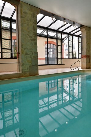 An indoor swimming pool with clear glass walls, stone pillars, and a view of buildings outside. - Hotel Castel Jeanson (Aÿ-Champagne)