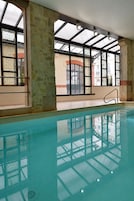 Una piscina interior con paredes de vidrio transparente, pilares de piedra y vistas a edificios exteriores.