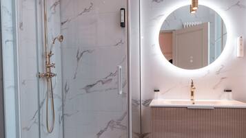Une salle de bain moderne dotée d’une douche à parois de verre, d’un meuble-lavabo avec un pare-éclaboussures en marbre et d’un miroir rond avec éclairage intégré.