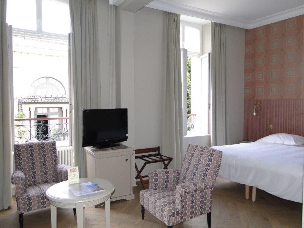 A hotel room with a bed, two patterned armchairs, a small table with a remote control, and a television on a stand.