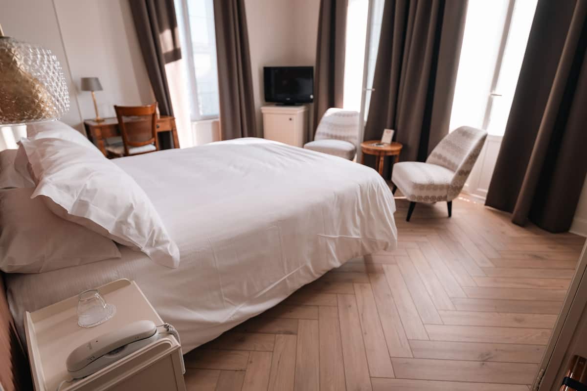 Ein Hotelzimmer mit einem großen Bett, einem Nachttisch mit Lampe, einem Sessel, einem Fernseher und Holzboden.