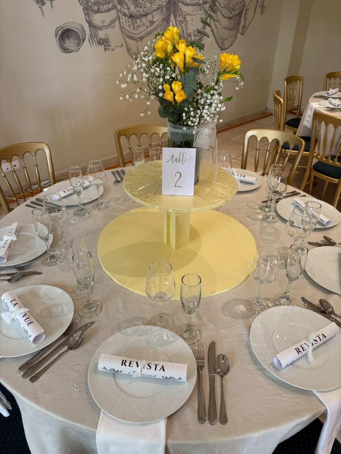 Ein runder Tisch ist für eine Mahlzeit gedeckt, mit einem Blumenstrauß aus gelben Blumen, einem Tischschild und Platzdeckern mit Namensschildern.