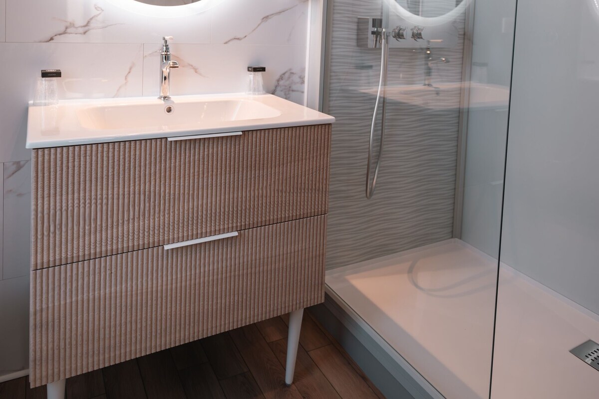 Un baño moderno con pared de mármol, un espejo redondo sobre el lavamanos y una ducha con paredes de vidrio.