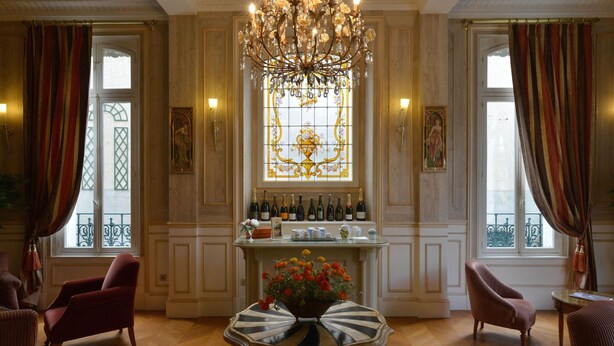 Una habitación con una araña, una chimenea con botellas de vino, una mesa con un arreglo floral y dos sillones.