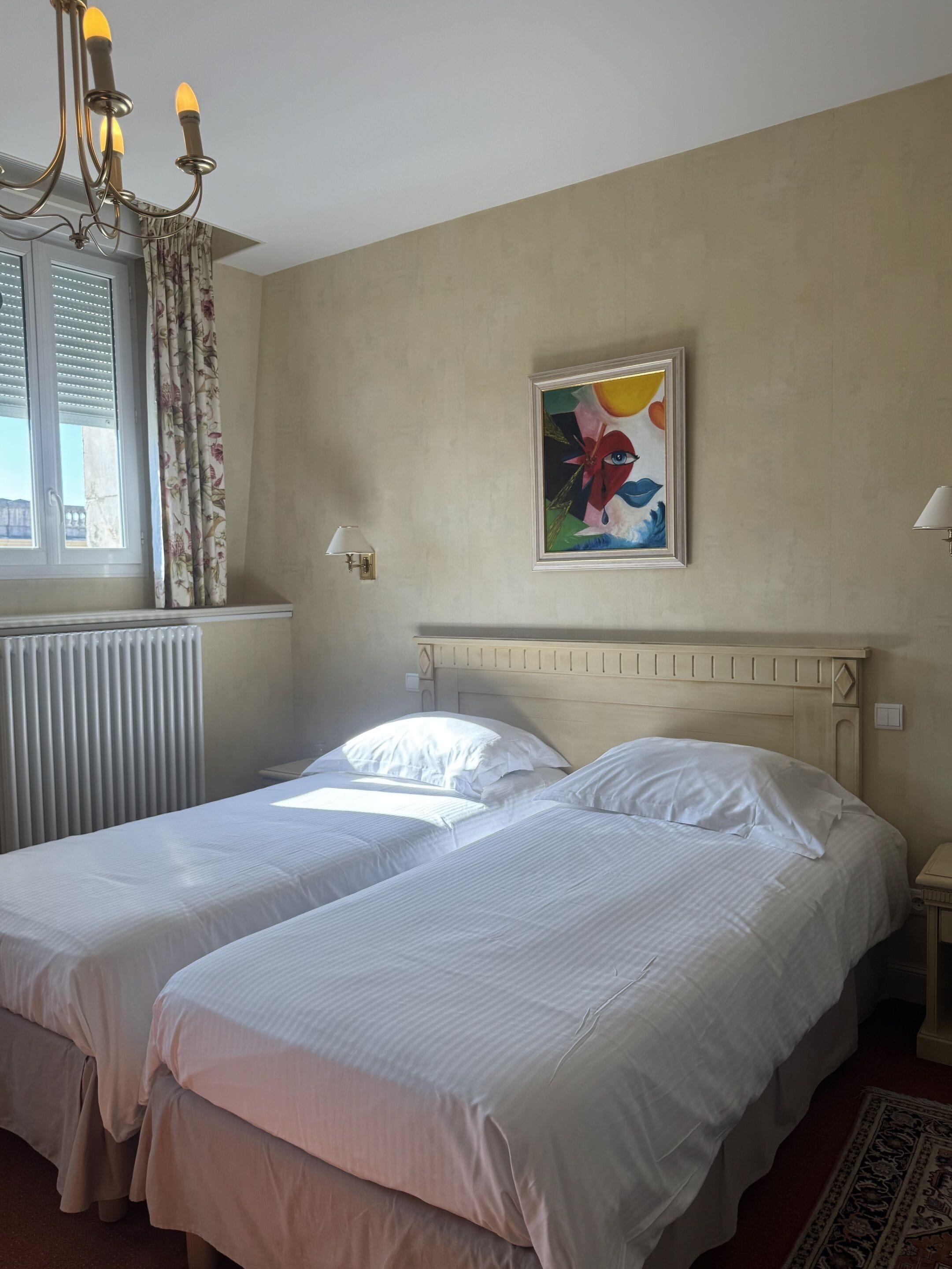 Ett hotellrum med två sängar, en kristallkrona, en tavla på väggen och ett fönster med gardiner.